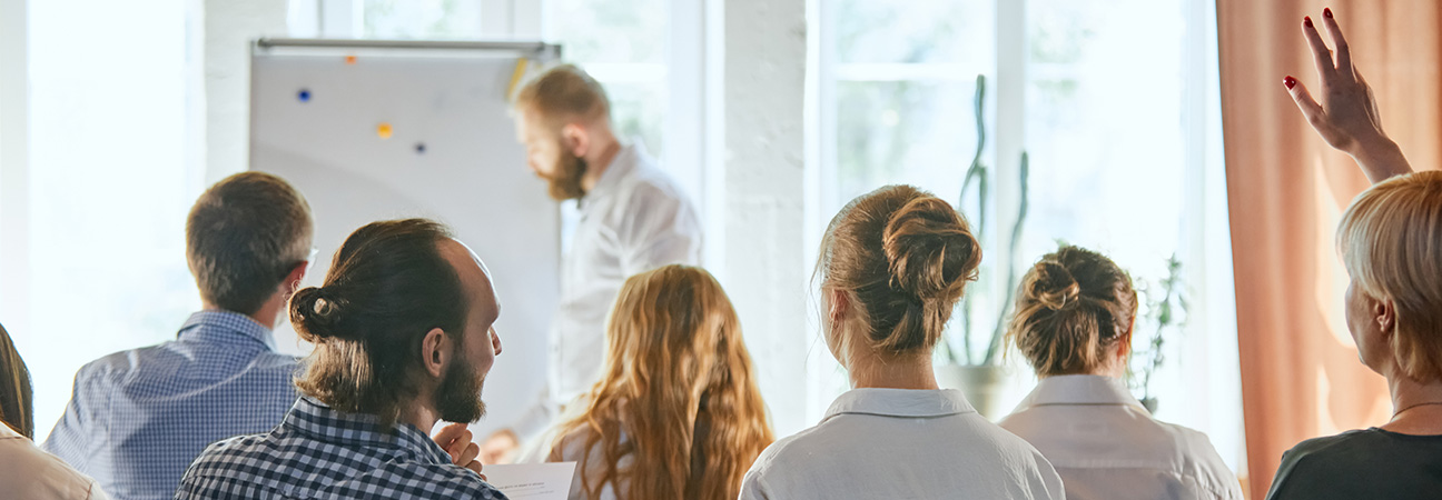Seminarteilnehmer sitzen in einem Raum und schauen auf eine Tafel. Ein Mann unterhält sich mit der Frau, die neben ihm sitzt. Eine Frau hält den Arm hoch. Ein Referent schreibt etwas auf die Tafel.